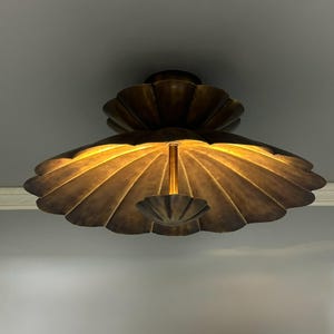 Könnte beinhalten: Eine bronzefarbene Deckenleuchte mit einem geschichteten, wellenförmigen Design. Die Leuchte hat einen zentralen Stiel und eine kleinere, umgekehrte Muschel am Boden. Das Licht strahlt einen warmen, goldenen Schein aus.