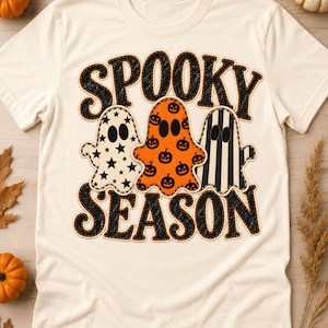 Może przedstawiać: Kremowy t-shirt z grafiką przedstawiającą czarny napis SPOOKY SEASON. Pomiędzy słowami znajdują się trzy kształty duchów: biały w czarne gwiazdki, pomarańczowy w czarne dynie oraz w czarno-białe pionowe paski.