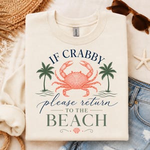 Puede incluir: Una sudadera de cuello redondo color crema con un gráfico de un cangrejo rojo centrado entre dos palmeras verdes. El texto dice IF CRABBY please return TO THE BEACH en una mezcla de fuentes azul marino y verde, con un pequeño icono de concha.