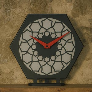 May include: A black, geometric wall clock with a decorative, light gray and black patterned face. The clock has red hands and sits on a wooden shelf. The clock is approximately 10 inches wide.