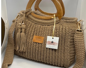 Puede incluir: Bolso de mano de crochet color beige con asas de madera y bandolera extraíble. El bolso presenta un patrón de puntadas texturizadas, una etiqueta de cuero y una borla decorativa. El bolso tiene un cierre y herrajes dorados. El bolso tiene una etiqueta que dice "V&L STYLE".