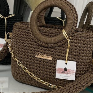 May include: A brown crocheted handbag with a circular handle and a gold chain strap. The bag features a gold "Fashion" emblem and a tag with the text "V&I style". The bag is part of a collection of crocheted bags.