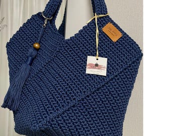 Puede incluir: Un bolso tote de ganchillo azul marino hecho a mano con un diseño geométrico. El bolso cuenta con asas resistentes, una borla decorativa con una cuenta de madera y una pequeña etiqueta que dice "V&I STYLE". El bolso está diseñado para transportar objetos.