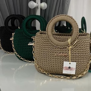 May include: Three crochet handbags in black, green, and beige, each with a circular handle and a gold chain strap. The bags have a woven texture and a small gold emblem. A tag hangs from the beige bag.