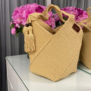 Puede incluir: Bolso tote de crochet beige con diseño geométrico. El bolso tiene un asa, una borla decorativa y una pequeña etiqueta de cuero marrón. El bolso se exhibe sobre una superficie reflectante, con un ramo de flores rosas en el fondo.