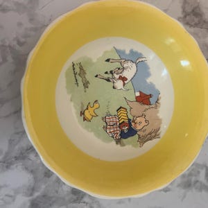 May include: A round ceramic bowl with a wide yellow rim and a white center. The interior features a colorful illustration of a pig playing an accordion, a yellow duck, and a white lamb on a green field, with the text Peter Rabbit printed nearby.