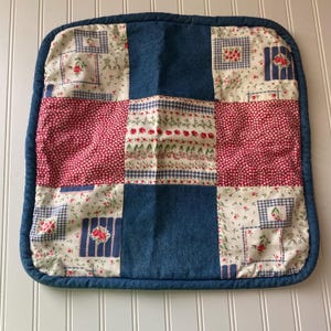May include: Square quilted pot holder with a patchwork design. The design includes denim blue squares, red polka dots, and floral patterns in white and blue. The edges are trimmed with a blue binding.