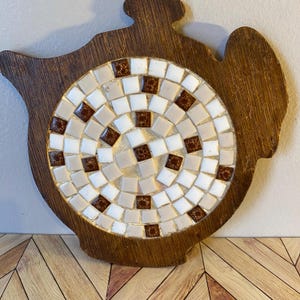 May include: A wooden teapot-shaped trivet. It has a circular mosaic design made of white, tan, and brown square tiles. The wooden base has a dark brown finish.