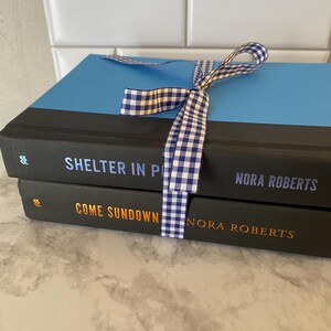 May include: Two hardcover books, "Shelter in Place" and "Come Sundown" by Nora Roberts, are tied together with a blue and white gingham ribbon. The top book has a blue cover, and the bottom book has an orange title.