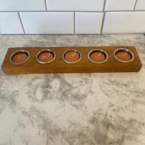 May include: A wooden candle holder with five metal-lined spaces, each holding a small, round, orange candle. The wooden base is rectangular and has a natural wood grain. The candles are unlit and set against a white tile background.