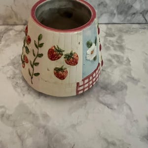 May include: A ceramic planter with a tapered shape, featuring a cream-colored base decorated with embossed red strawberries, green vines, a white flower on a light blue panel, and a red and white checkered pattern along the bottom edge.
