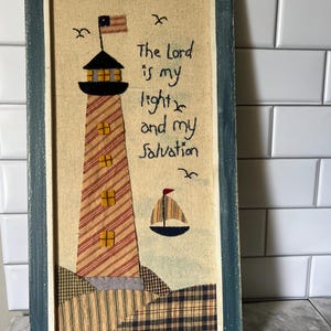 May include: A rectangular wall art piece featuring a fabric applique of a lighthouse with red and white stripes, a small sailboat, and embroidered text that reads, The Lord is my light and my salvation, all enclosed in a distressed blue wooden frame.