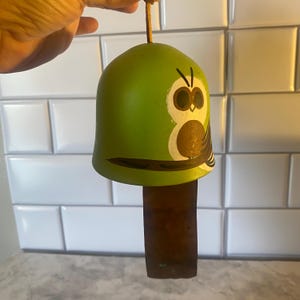 May include: A green bell-shaped wind chime with a painted owl design. The owl has large brown eyes and is perched on a branch. The chime has a brown, rectangular clapper and a string for hanging.