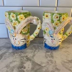 May include: Two ceramic mugs with a bunny rabbit design. The mugs are white with yellow flowers and green leaves. The bunny face is on the front, with a blue bow tie. The handles are shaped like bunny ears.