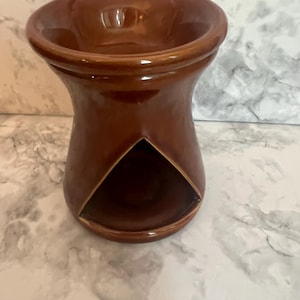 May include: A glossy, dark brown ceramic wax warmer featuring a wide, circular basin at the top for wax melts and a large, triangular-shaped opening at the base designed to accommodate a small tea light candle for heating purposes.