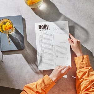 May include: A "Daily Planner" sheet with a structured layout is held by a person wearing an orange shirt. A blue notebook with a glass of orange-infused beverage and a gold straw sits to the left. The scene is set on a light gray surface.