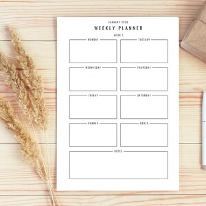 Puede incluir: Un planificador semanal blanco para enero de 2026 con secciones para cada día de la semana, objetivos y notas. Un bolígrafo, un regalo envuelto y plantas secas están sobre una superficie de madera clara.