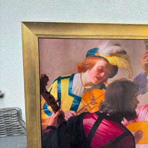 May include: A framed print of a painting depicting two figures, one with reddish hair wearing a yellow and blue outfit with a feathered cap, holding a violin, and another figure in a dark red garment with a black cape, both engaged in music.