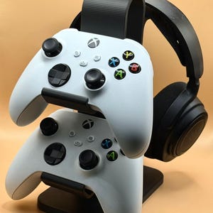 May include: A black gaming headset and two white Xbox controllers are displayed on a black stand. The controllers are stacked, and the headset is positioned to the right. The controllers have black joysticks and coloured buttons.