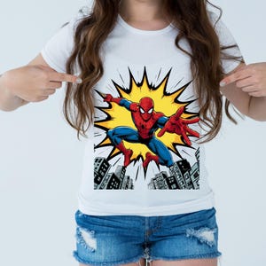 May include: White t-shirt featuring a colorful graphic of Spider-Man in action. The superhero is depicted in his classic red and blue suit, set against a yellow burst and a cityscape background. The design is a vibrant and eye-catching.