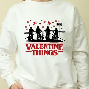 Puede incluir: Sudadera blanca con un diseño rojo y negro. El diseño presenta siluetas de personas en bicicleta, cupidos, corazones y el texto "VALENTINE THINGS" y "WELCOME HAWKINS".
