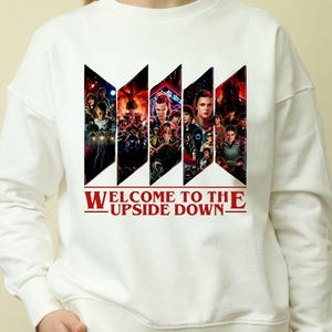 Puede incluir: Sudadera blanca con un diseño gráfico con personajes de una serie de ciencia ficción. El diseño está segmentado en formas geométricas negras con imágenes de los personajes. El texto "WELCOME TO THE UPSIDE DOWN" está impreso en rojo.