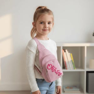 May include: A pink corduroy sling bag with the name "Olivia" embroidered in a cursive font. The bag has a zippered front pocket and a matching strap. The bag is worn by a young girl wearing a white sweater and blue jeans.