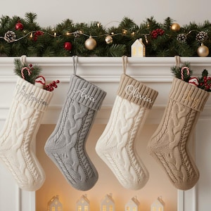 Könnte beinhalten: Vier Zopfstrick-Weihnachtsstrümpfe in Creme, Grau und Beige hängen an einem weißen Kaminsims. Jeder Strumpf hat einen personalisierten Namen auf der Manschette. Die Strümpfe sind mit festlichen Dekorationen gefüllt, darunter Zuckerstangen und Grünzeug.