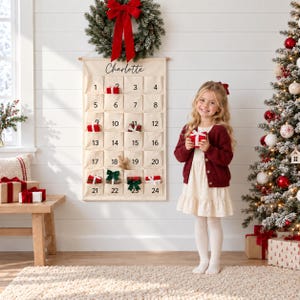 Könnte beinhalten: Ein cremefarbener Stoff-Adventskalender mit vierundzwanzig nummerierten Taschen und dem aufgedruckten Namen Charlotte. Der Kalender hängt an einer Wand unter einem grünen Kranz mit einer großen roten Schleife und zeigt kleine Geschenke in den Taschen.