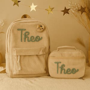 May include: A beige corduroy backpack and matching lunch bag, both with the name "Theo" embroidered in green. The backpack has a front pocket and a cat-shaped patch. A gold star garland hangs in the background.