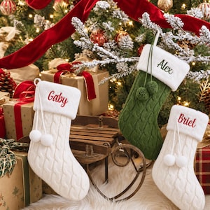 Op de afbeelding: Drie kerstsokken van kabelgebreid materiaal met witte randen van imitatiebont. Twee sokken zijn wit met de in rood geborduurde namen Gaby en Briel, en één is olijfgroen met de naam Alex in donkergroen geborduurd. Elke sok heeft twee pompons.