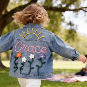 May include: A light blue denim jacket with the name "Grace" embroidered in pink script across the back. The jacket features a yellow sun design and colourful flower appliqués. The jacket is designed for children.
