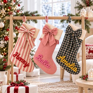 May include: Three Christmas stockings hanging on a wooden stand. The first is red and white striped with a bow and the name Henry. The second is solid pink with a ruffled trim, a bow, and the name Bella. The third is green and black plaid with a bow and the name Amelia.