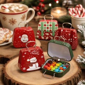 May include: A collection of small, suitcase-shaped metal tins featuring festive holiday designs, including Santa Claus, a snowman with the text Merry Christmas, and geometric patterns. One green tin is open, revealing colourful sweets inside.