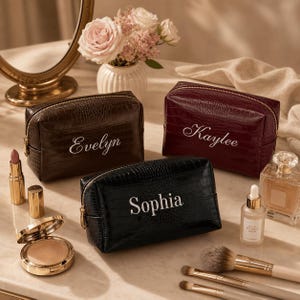 May include: Three rectangular cosmetic bags with a crocodile-embossed texture in dark brown, burgundy, and black. Each bag features a gold zip and a white embroidered name: Evelyn, Kaylee, and Sophia. The bags are arranged with makeup items.