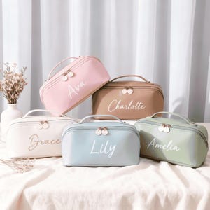 May include: Five pastel-coloured cosmetic bags, each with a handle, zip, and a name printed in cursive. The bags are pink, tan, white, blue, and green. They are arranged on a white surface. Each bag has a gold-coloured zip.