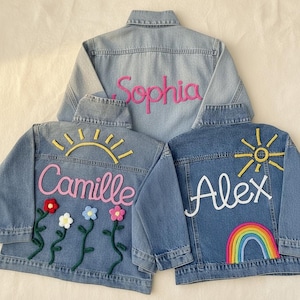May include: Three denim jackets with embroidered details. One jacket has "Sophia" in pink script, another has "Camille" with flowers and a sun, and the third has "Alex" with a rainbow and sun.