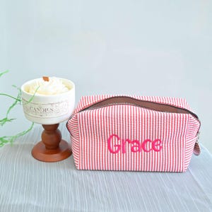 Peut inclure: Une trousse de maquillage à rayures rouges et blanches avec le nom "Grace" brodé en rose. Le sac a une fermeture éclair et un intérieur marron. Une bougie blanche dans un support décoratif est en arrière-plan.