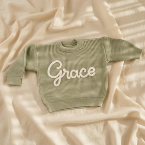 May include: A sage green knitted sweater with the name "Grace" embroidered in white script across the front. The sweater has a ribbed texture and long sleeves. The garment is displayed on a cream-coloured fabric.