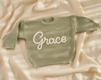 May include: A sage green knit sweater with the name "Grace" embroidered in white script across the front. The sweater has a ribbed texture and long sleeves. The garment is displayed on a cream-colored fabric.