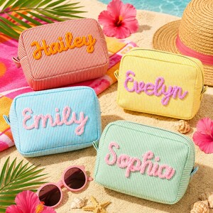 Puede incluir: Cuatro estuches rectangulares con cremallera en rojo, amarillo, azul claro y verde menta, cada uno con un patrón de rayas blancas finas. Los estuches están decorados con letras de lana en relieve en cursiva que forman los nombres Hailey, Evelyn, Emily y Sophia.
