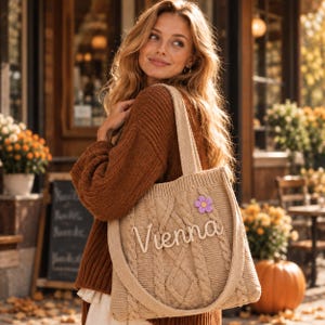 Puede incluir: Un bolso tote de punto trenzado en color beige con el nombre Vienna en letras cursivas blancas y un pequeño adorno de flor morada. El bolso cuenta con dos asas de punto a juego y un patrón de tejido grueso y texturizado en toda la superficie.