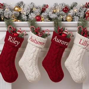 Puede incluir: Cuatro calcetines navideños de punto trenzado, dos rojos y dos blancos, cuelgan de una repisa blanca. Cada calcetín está personalizado con un nombre bordado en blanco: Riley, Autumn, Aurora y Lilly. Los calcetines están llenos de bastones de caramelo y adornos.