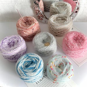 May include: A selection of colourful yarn balls in shades of pink, purple, blue, and grey. Some balls have speckled or variegated patterns. A needle is inserted into one of the pink yarn balls. The text "TODAY" is partially visible.