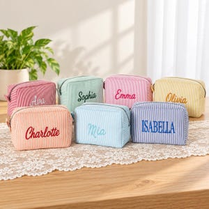 May include: A collection of seven rectangular zipped pouches made of striped fabric in various pastel colours. Each pouch features a unique name embroidered on the front in a contrasting thread colour, including Ava, Sophia, Emma, Olivia, Charlotte, Mia, and Isabella.