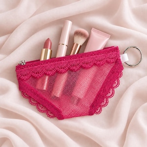 May include: A fuchsia pink mesh zipped pouch with a scalloped lace trim along the top edge and a silver metal keyring attached to the side. The pouch contains a lipstick, a cylindrical tube, a makeup brush, and a small squeeze tube of cream.