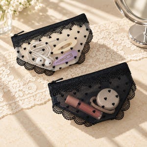 May include: Two semi-transparent zippered pouches featuring a black lace trim and a pattern of small black hearts. One pouch is cream-coloured containing white wired earbuds and two hair clips, while the other is black containing a lip gloss and hair tie.