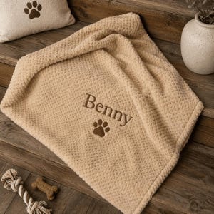 Peut inclure: Une couverture beige à texture gaufrée présentant le nom Benny brodé en fil marron foncé, accompagné d'une icône d'empreinte de patte marron foncé. Le tissu doux possède un motif en relief et est plié pour afficher le texte personnalisé.