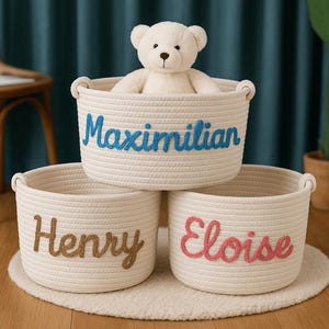 Puede incluir: Tres cestas de cuerda color crema con nombres bordados: Maximilian, Henry y Eloise. Un osito de peluche blanco está sentado en la cesta superior. Las cestas están sobre una alfombra redonda blanca.