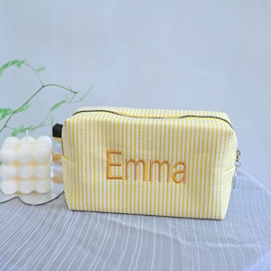 Op de afbeelding: Een geel en wit gestreept cosmetica tasje met de naam "Emma" geborduurd in goudkleurig garen. De tas heeft een zwarte rits en een zilveren ritstrekker. De tas is rechthoekig en ligt op een lichtgrijze ondergrond.